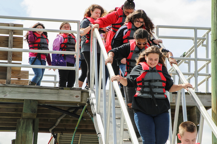 Middle school student walk down to a dock wearing life jackets.
