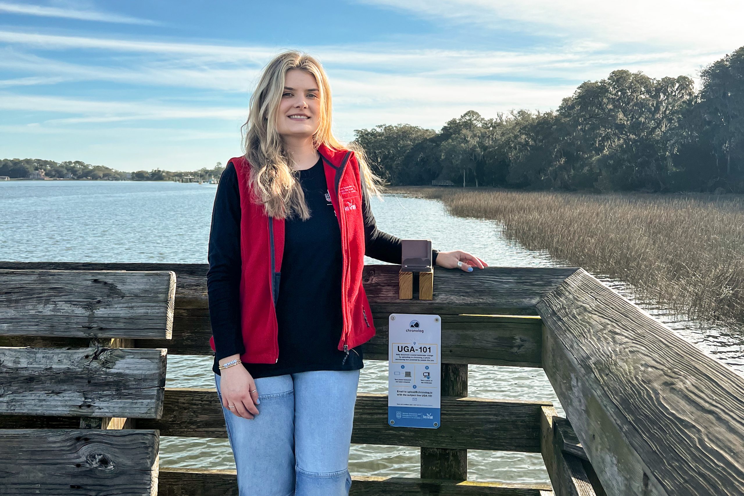 a young woman with blonde hair stands on a wooden observation dock overlooking a marsh and waterway