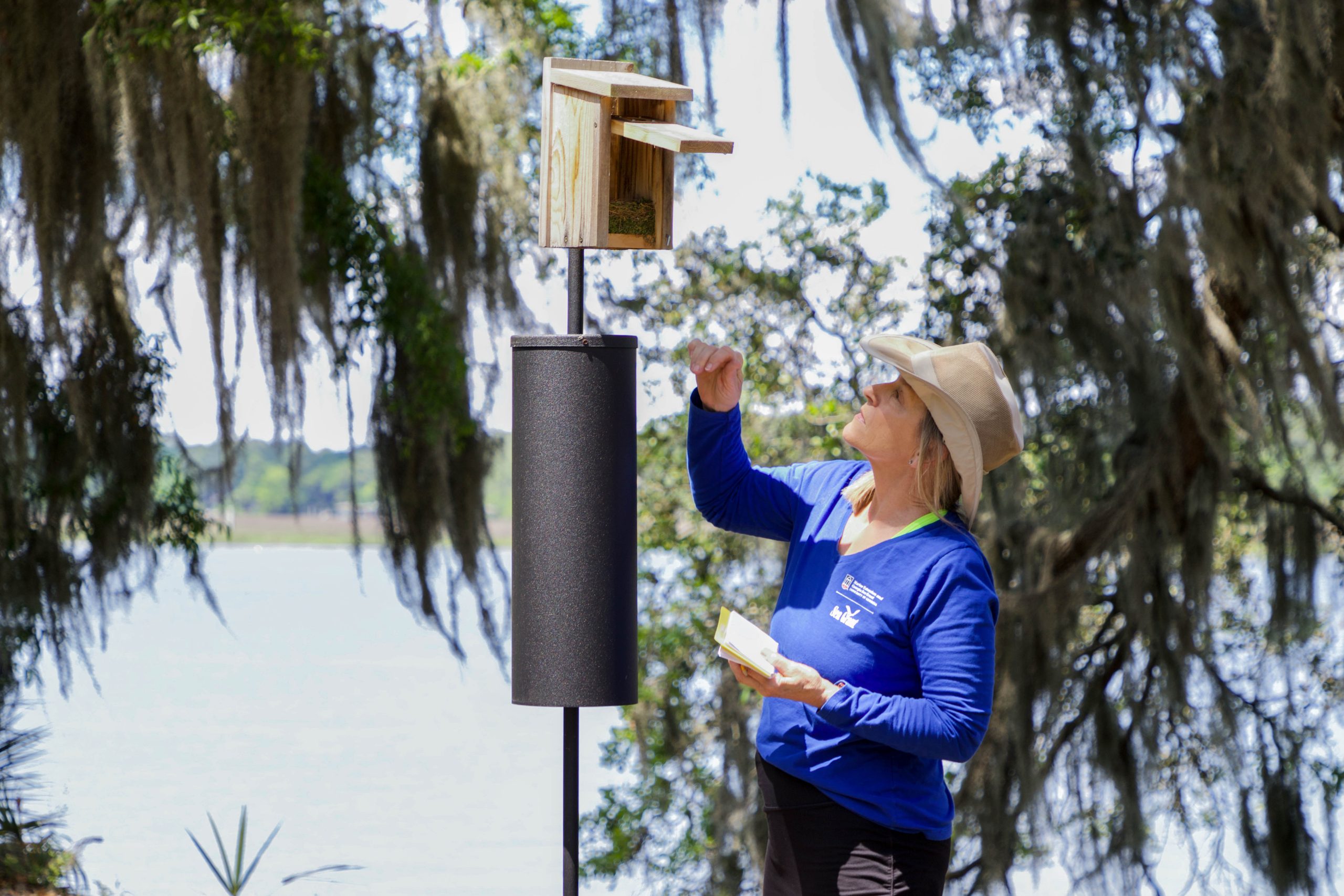 a woman wearing a blue shirt and a brimmed hat holds a notebook in one hand and reaches toward an open bird nesting box with the other