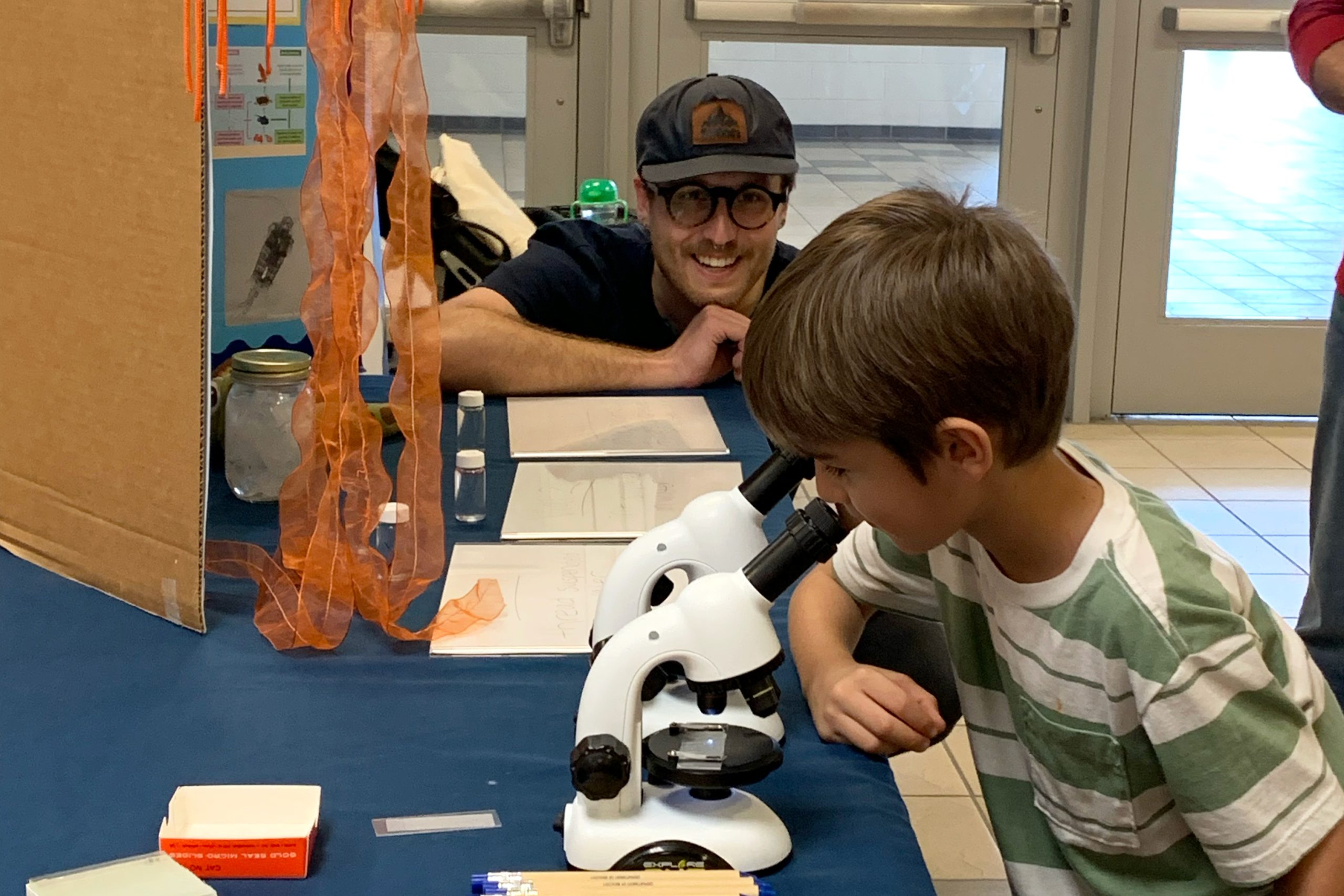 a man smiles as he watches a child look through a microscope