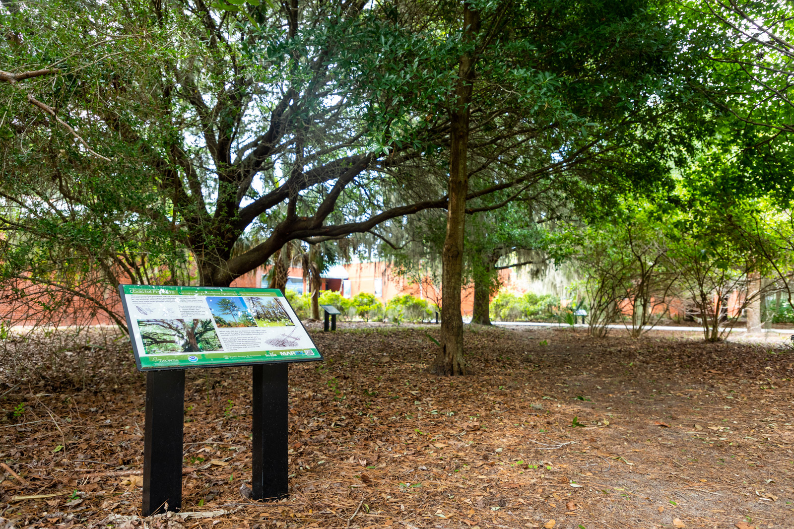 An educational interpretive sign stands along a nature trail shaded by dense trees, with the red brick exterior of the aquarium visible in the background.