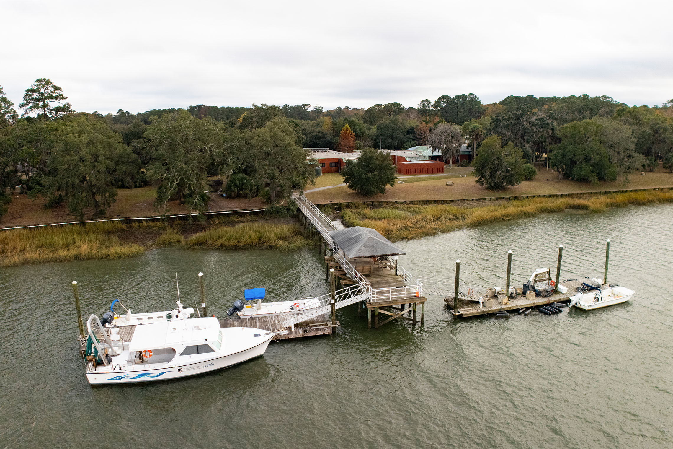 Aerial view of several white boats moored at a dock along a wooded shoreline at the UGA Marine Education Center and Aquarium