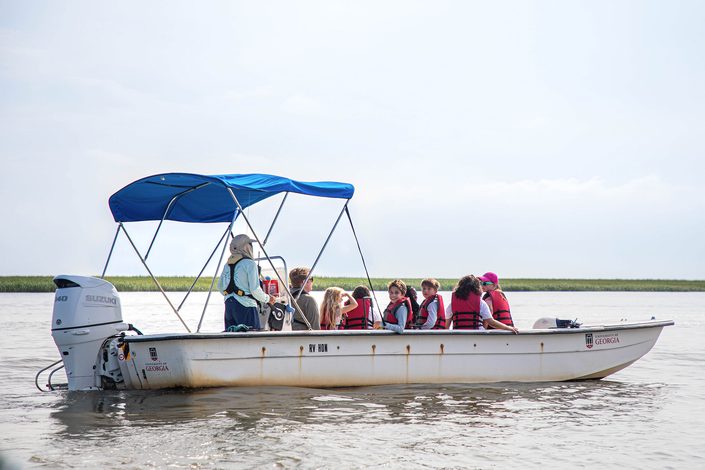 A white boat with a blue canopy, filled with students wearing life vests, cruising along the marshy coastline