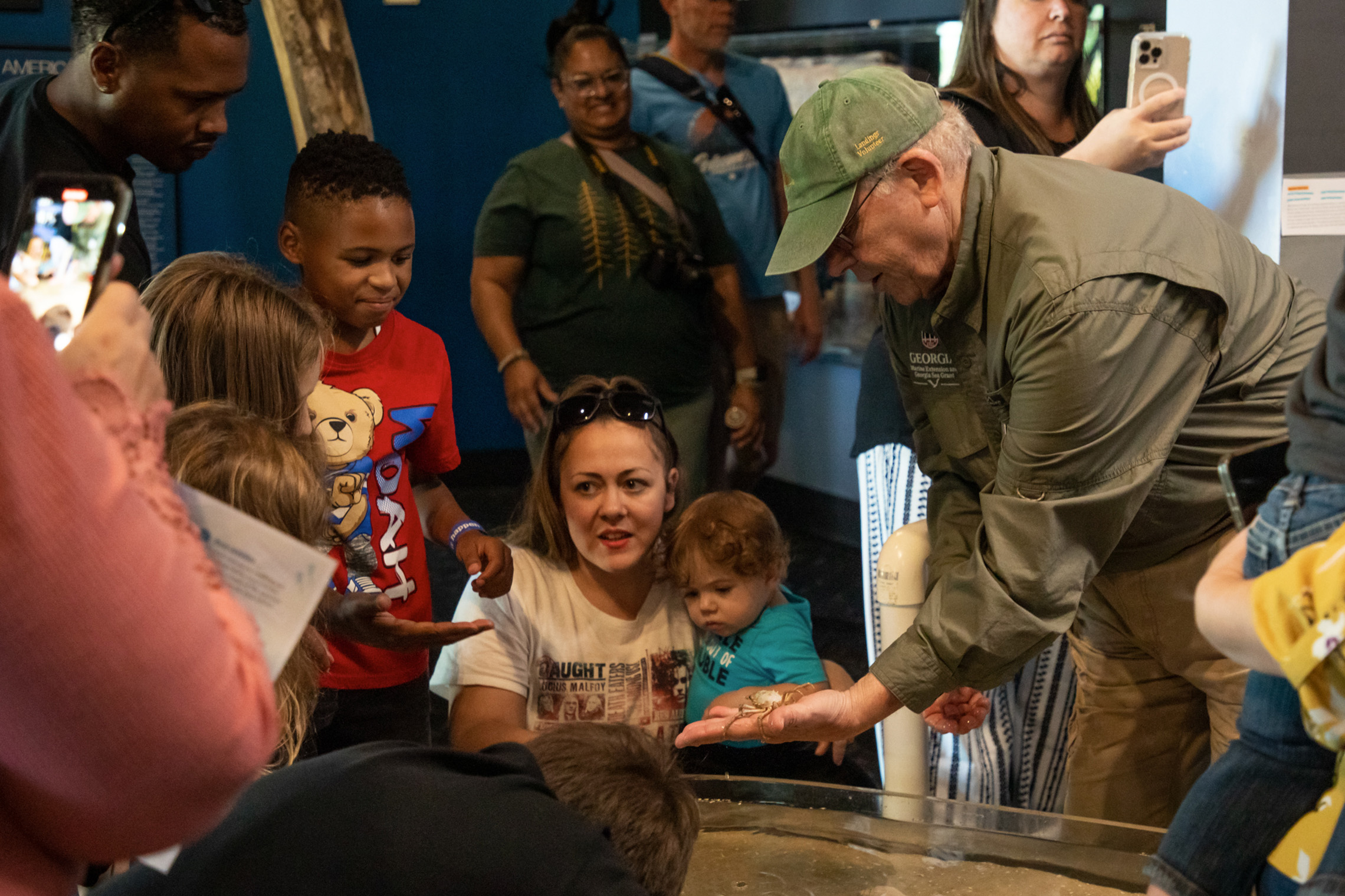 A group of people gather around a touch tank in an aquarium. A man in a green cap holds a small spider crab, as onlookers, including children, watch with interest.
