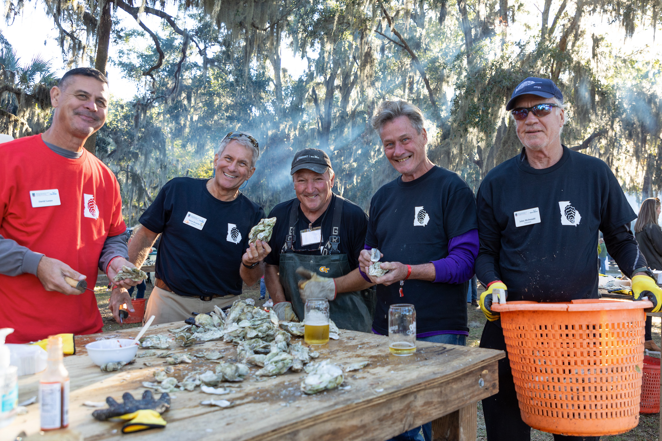 Five men smiling while shucking oysters at an outdoor event. They're gathered around a wooden table, surrounded by trees and Spanish moss.
