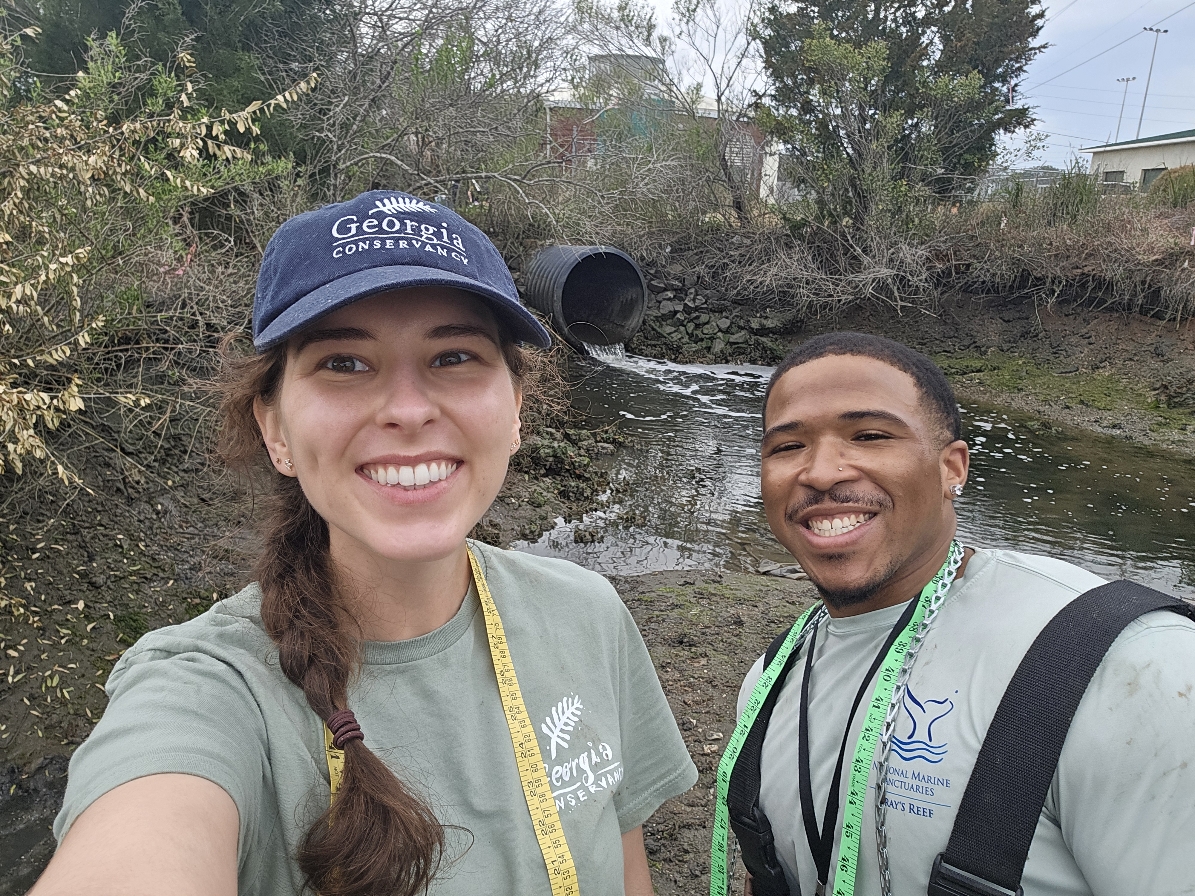 a young woman and man stand next to each other smiling with measuring tapes around their necks with a creek in the background