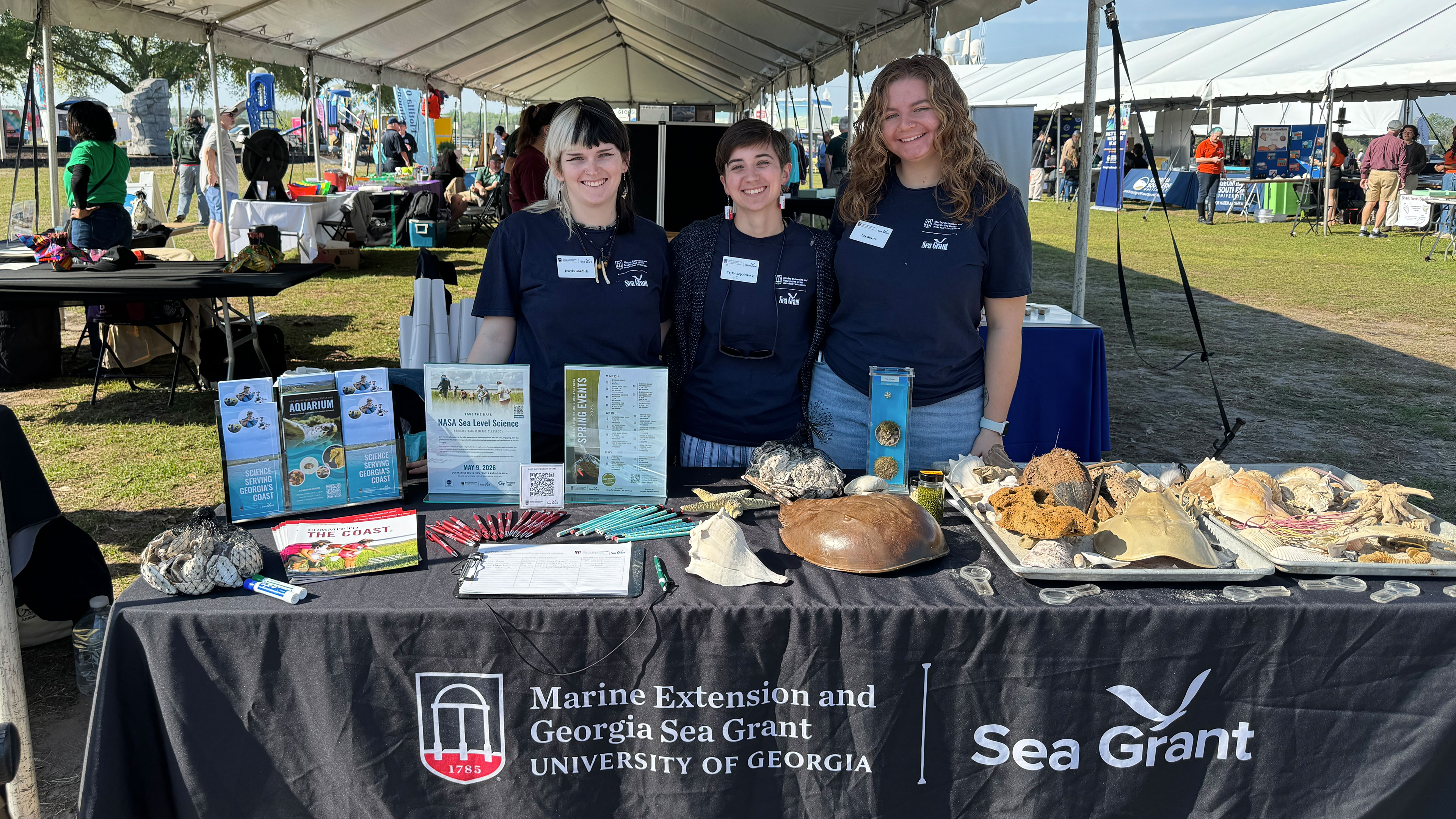 three young adults smile at event with a table in front of them full of outreach materials and shells