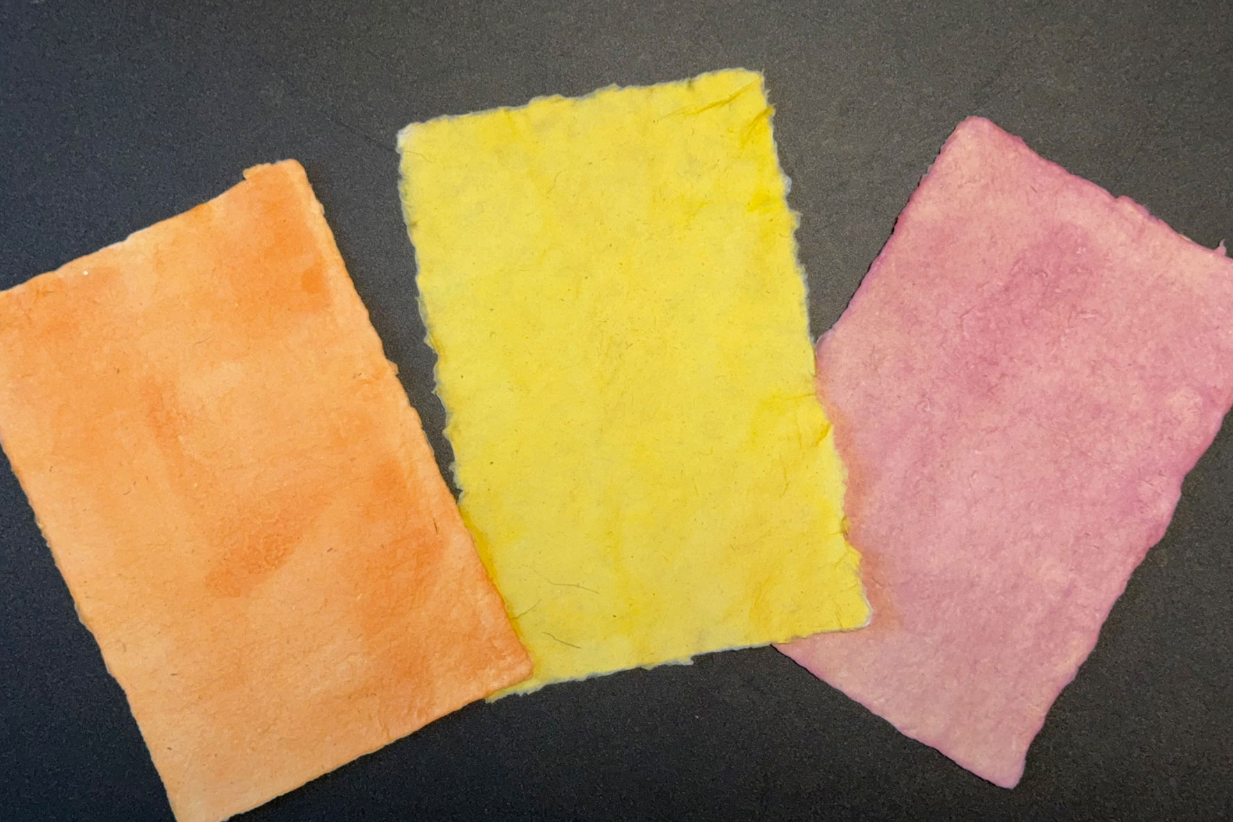 three pieces of handmade paper samples in orange, yellow, and pink