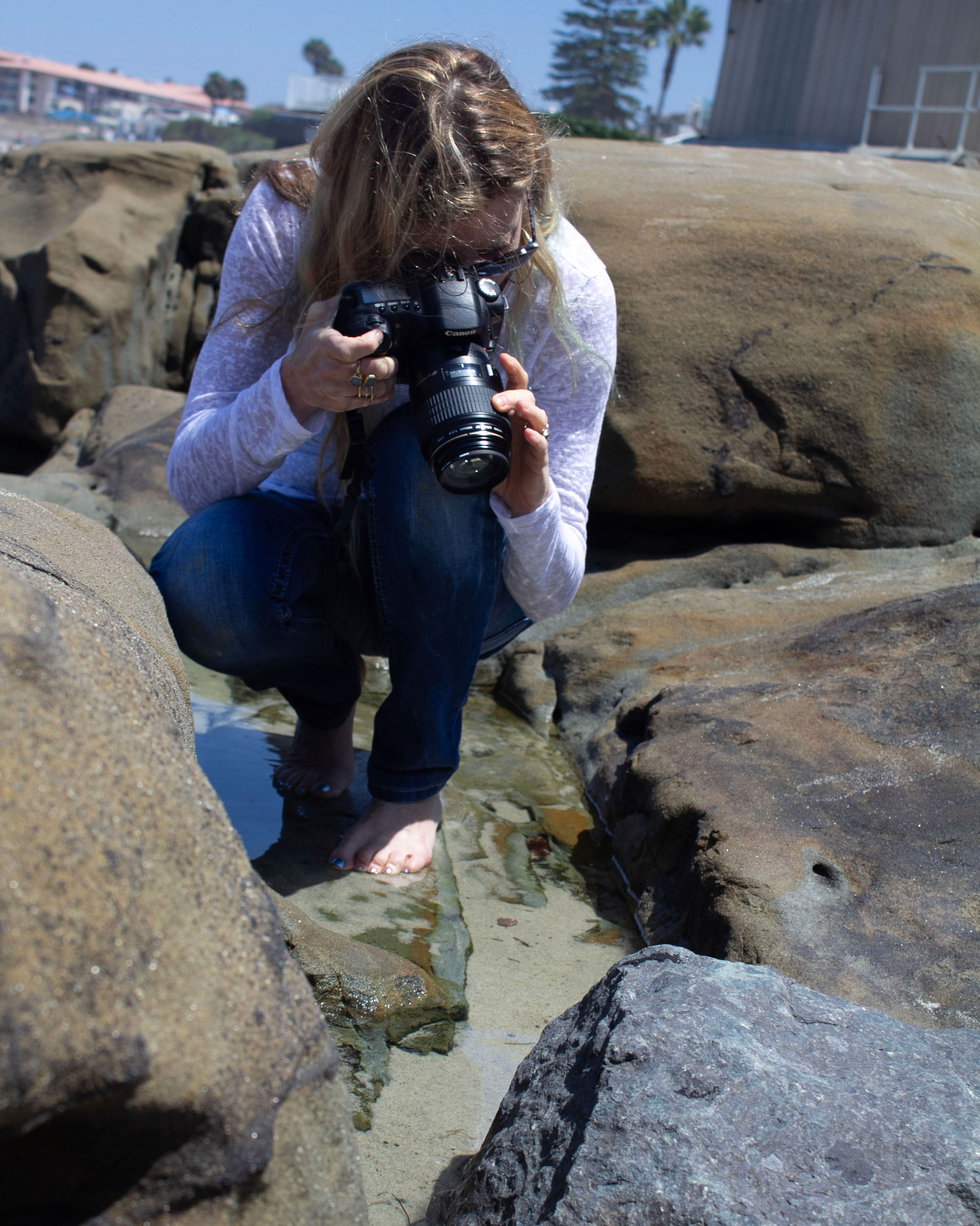 a woman crouches near the ocean on rocks and takes a photograph with a camera