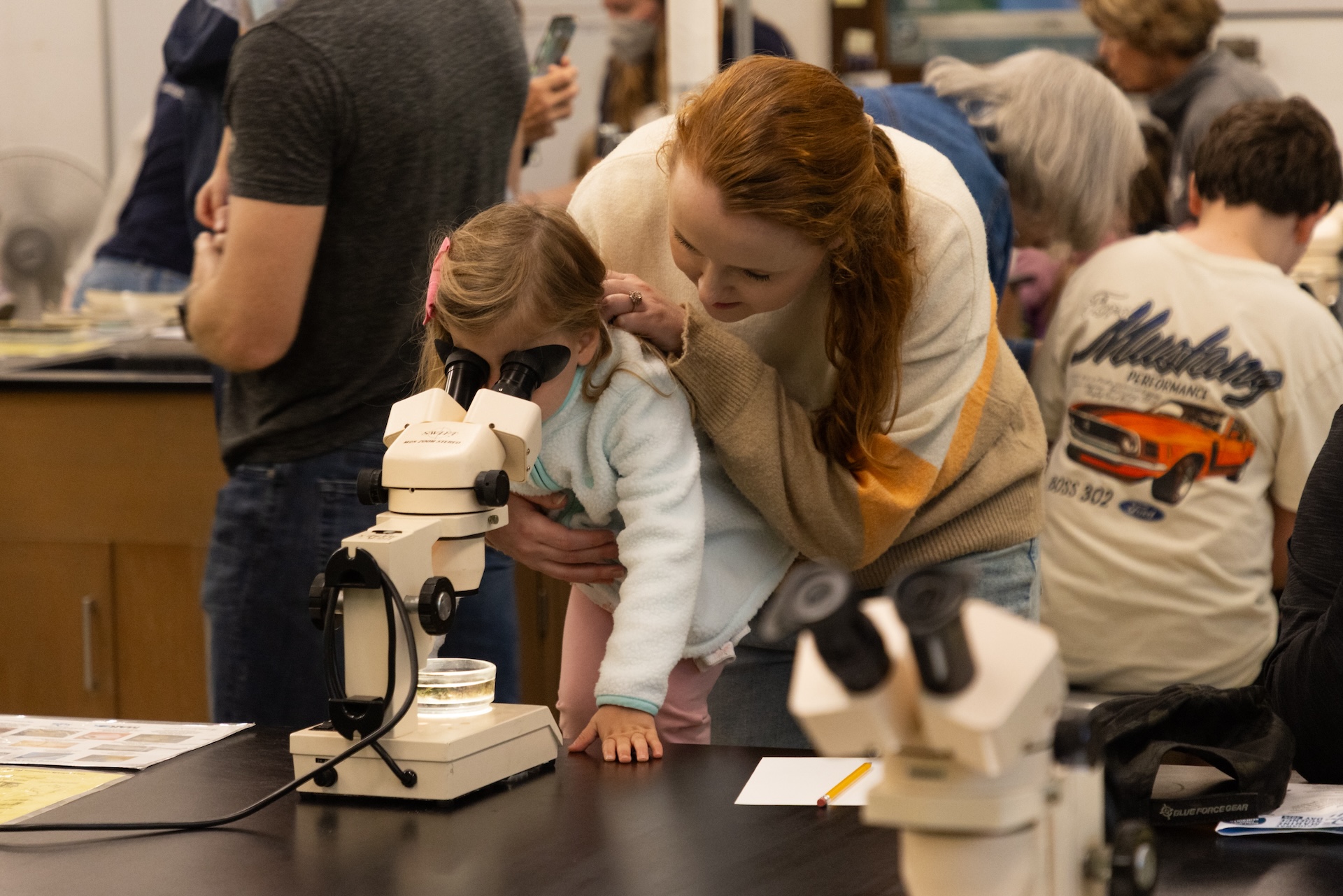 Woman and little girl look through microscope in classroom setting.