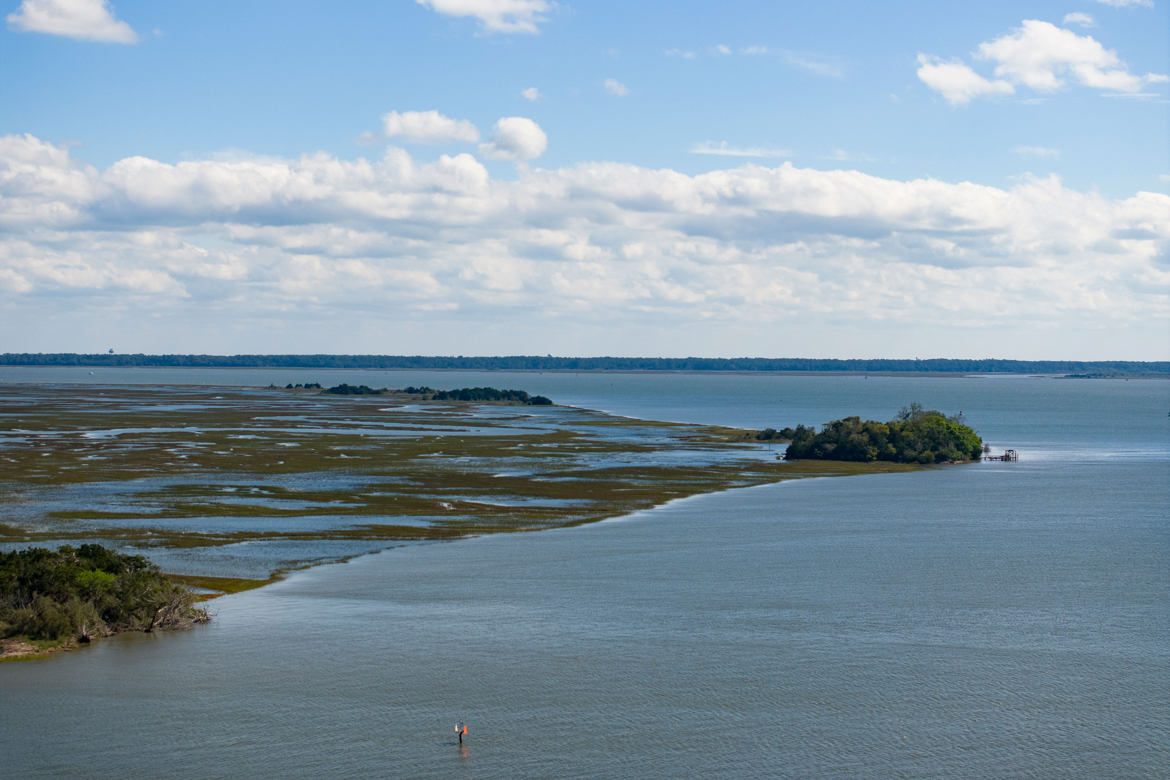 A tidal marsh and a small island surrounded by water under a cloudy blue sky.
